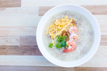 Boiled rice with pork was added on top with chopped omelet, boiled shrimp and coriander as a a breakfast meal in the morning in Thai style while that bowl put on wooden background.