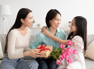 Family holidays. Little girl with her mom congratulating granny with birthday, presenting flowers and gift at home