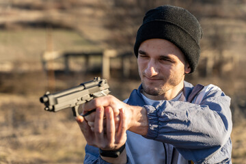 Close up portrait of adult young caucasian man wearing black cap hat holding a gun pointing and aiming in sunny day outdoor - criminal or gangster terrorism murderer with firearm concept copy space