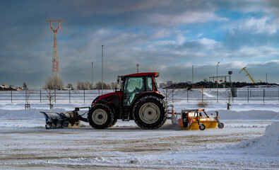 Red  tractor is cleaning car parking zone from snow with snow plow and rotating brush.