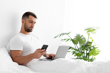Happy caucasian attractive young guy in white t-shirt typing blog on computer and look at phone