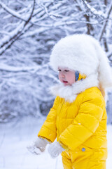 Portrait of a frozen little three-year-old girl in a yellow jumpsuit and a white fluffy hat. Close-up of a pretty caucasian girl in winter clothes in a snowy park. The child is very cold.