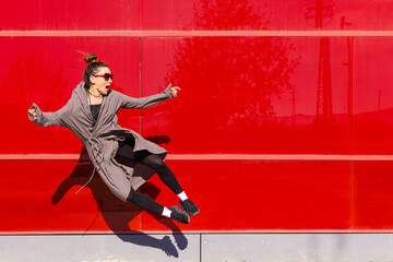 middle-aged woman dressed in casual alternative clothing, jumping in the street in front of a bright red wall