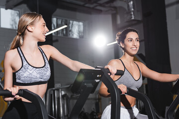 Cheerful interracial women training on elliptical trainers in sports center.