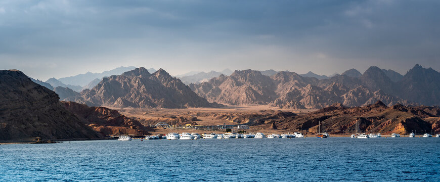 Yachts and tourist boats moored in a sea harbor of Sharm El-Sheikh, view on a coast with panorama of mountains. Mountain landscape with tourist cruise boats near the Ras-Mohammed Reserve.
