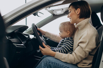 Woman with her little son holding steering wheel in a car in a car showroom