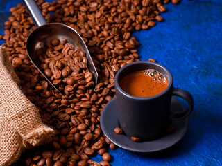 Dark grey espresso mug, coffee beans scattered on blue concrete background table from rustic sack, metal scoop. Coffee shop, food, roaster, morning drink concept. Close up