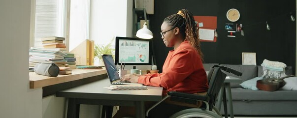 Zoom in shot of young African American woman with disability sitting in wheelchair at desk, using laptop and taking notes while working online from home - Powered by Adobe