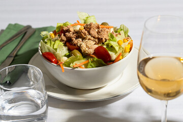 Tuna salad with lettuce, tomatos, carrot, corn in the white bowl served on the white tableware in restaurant with glass water , white wine and silver cutlery