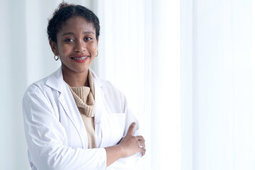 Young African American female doctor with charming smile wears  lab coat and holds tablet in hand, stand with arms crossed near the window