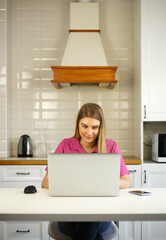 Fototapeta na wymiar Freelancer woman working at home. Beautiful blonde female using modern laptop computer for distant work during lockdown. Cheerful entrepreneur person typing on notebook pc in home kitchen
