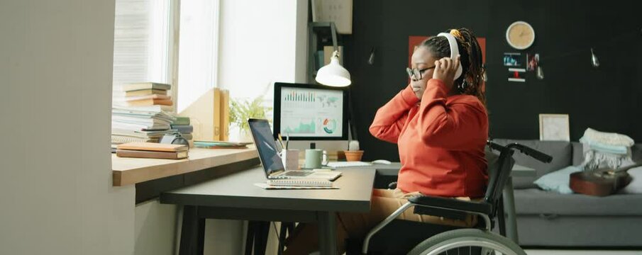 Zoom In Shot Of Young African American Woman With Disability Sitting In Wheelchair At Home Workplace, Putting On Wireless Headphones, Enjoying Music And Dancing