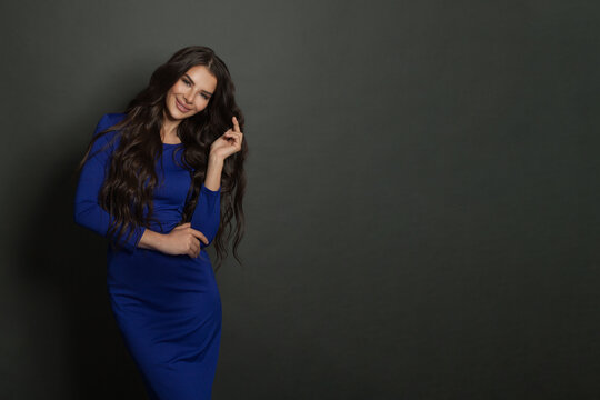 Happy young woman with long brown hair wearing blue dress smiling and posing against black background with copy space