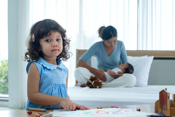 Little girl lookin at camera, baby sister and her mother on the bed in the back, girl in foreground is clear and sharp and mom and baby in back are blurred