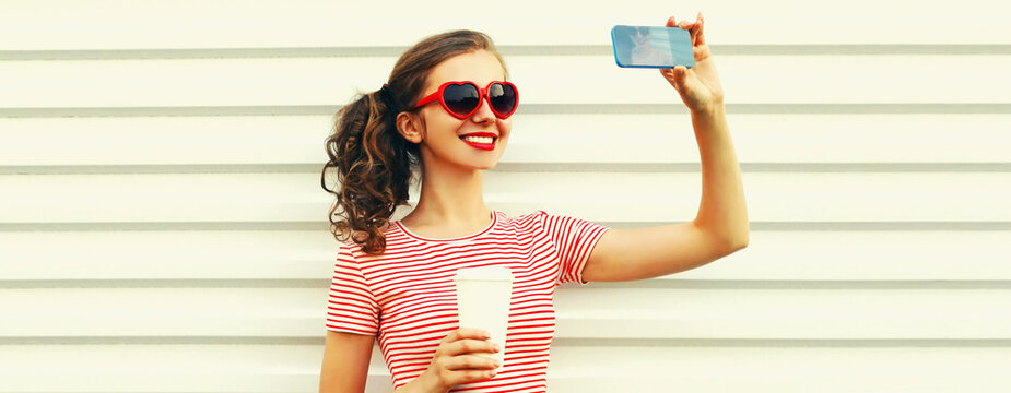 Portrait of happy smiling young woman taking selfie by phone with coffee cup wearing red heart shaped sunglasses on white background