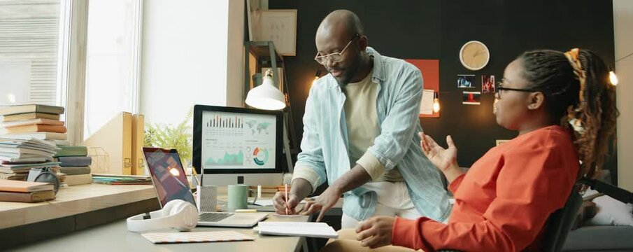 Zoom out shot of African American woman with disability sitting in wheelchair at desk and discussing business project with male colleague as he taking notes while working together in home office
