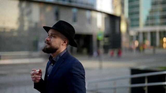 a bearded and mustachioed man in a hat and a plaid jacket holds a cigarette in hand and walks against the background of a blurred city street. side view. the camera is moving
