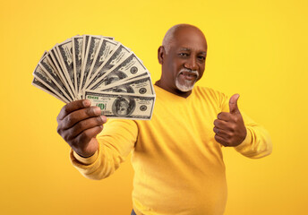 Happy senior black man holding fan of money and showing thumb up gesture on orange background,...