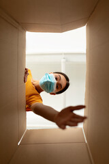 Young asian woman in medical mask unboxing cardboard parcel, trying to get something, view from the inside
