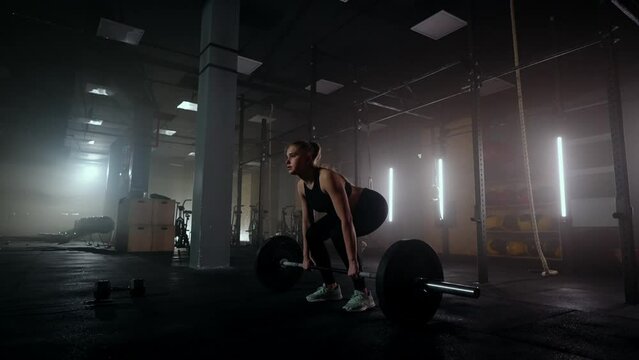 Slow motion: A woman lifts a barbell in a dark gym with a lot of weight. A strong young woman lifts weights for a workout