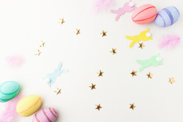 Festive background with paper rabbits and colorful eggs and golden confetti on a white wooden background, top view. Easter card with copy space and decorations.