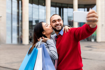 Affectionate young Asian woman kissing her boyfriend, holding shopper bags, expressing love near...