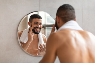 Happy guy looking in the mirror applying cream on face