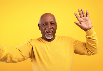 Happy senior black man taking selfie, waving at camera on orange studio background