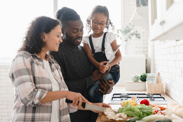 Cheerful african-american family of three, parents with daughter cooking together at home kitchen,...