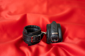 Accessories for bdsm on a red satin fabric background. Black color fetish leather handcuffs. Valentine's Day.
