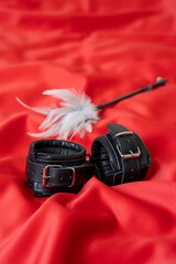 Accessories for bdsm on a red satin fabric background. Black color fetish leather handcuffs and feather tickler . Valentine's Day.
