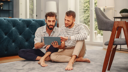 Two Stylish Young Adult Men Sitting on Living Room Floor at Home and Using Tablet Computer. Friends...