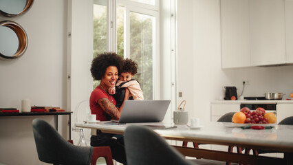Beautiful Young Mom Holding Her Lovely Baby Son, Using Laptop Computer in Living Room in Apartment. Mother is Working from Home, Online Shopping, Watching Social Media or Writing Emails.