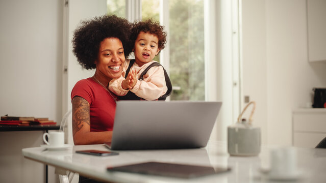 Beautiful Young Mom Holding Her Lovely Baby Son, Using Laptop Computer in Living Room in Apartment. Mother is Working from Home, Online Shopping, Watching Social Media or Writing Emails.