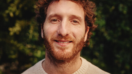 Close Up Portrait of a Happy Young Adult Male with, Brown Eyes, Curly Ginger Hair and Beard Posing for Camera. Handsome Diverse Caucasian Male Smiling on Green Nature Background. - Powered by Adobe