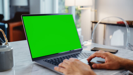 Fototapeta na wymiar Green Screen Mock Up Display on a Laptop Computer. Close Up on Person's Hands Working from Modern Home, Using Touch Pad, Scrolling Content. Smartphone Lies Next to Computer.