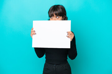 Young latin woman isolated on blue background holding an empty placard and hiding behind it