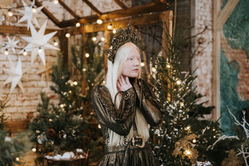 young albino woman with blue eyes and long white hair in beautiful green dress and crown stands in loft room decorated with wooden greenhouse and Christmas trees with twinkle lights, diverse people