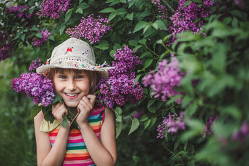 Laughing European brunette girl in a beige hat and multicolored striped dress holds lilac flowers in her hands