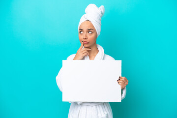 Young woman in a bathrobe with towel isolated on blue background holding an empty placard and thinking