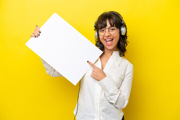 Telemarketer latin woman working with a headset isolated on yellow background holding an empty placard with happy expression and pointing it