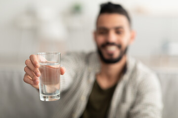Young Arab man showing glass of fresh clear water at camera at home, selective focus. Wellness...