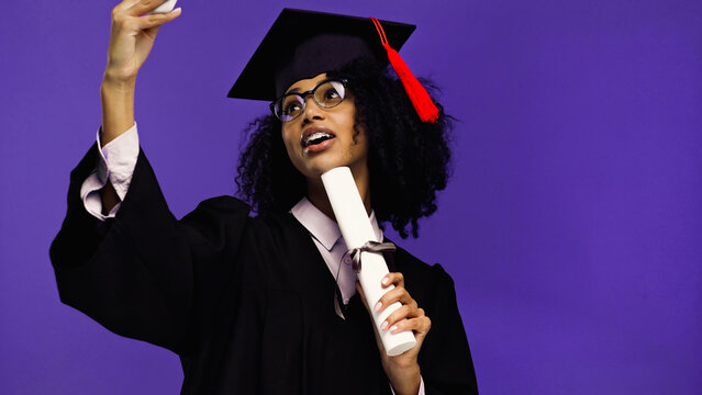 happy african american student with braces in graduation cap and gown taking selfie with rolled diploma isolated on purple.