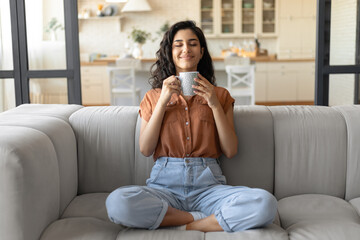 Pretty young Caucasian lady sitting cross legged on couch, smelling fresh aromatic coffee with...