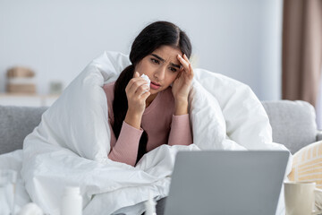 Sad young indian lady wrapped in blanket with napkin suffer from headache and fever, on sofa