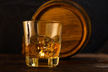 A glass of whiskey and ice stands on oak planks against the background of an oak barrel. Close-up. Whiskey from a home distillery.