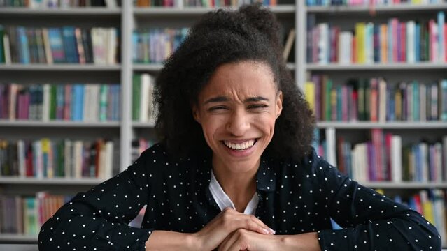 Cheerful African American female student with curly hair, sitting in the library, laughing during video communication, heard something funny, the girl is fooling around, smiling