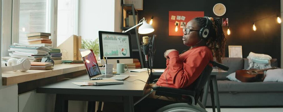 Young African American woman with disability sitting in wheelchair at desk with laptop and computer and speaking in microphone while recording business podcast in home studio
