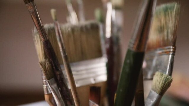 Closeup view of young female painter hand grabbing a paint brush from a container with many paintbrushes of different sizes and styles.
