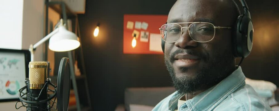 Portrait of African American male podcaster in headphones posing for camera while sitting in home recording studio